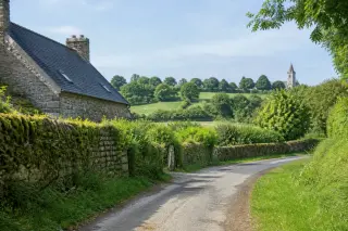 Chemin de campagne bordé de murets en pierre, ancienne ferme en granit, clocher d’église visible au loin dans le Cotentin sous une lumière douce
