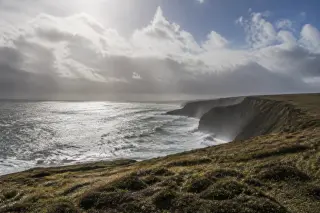Côte normande escarpée dans la Hague avec falaises, landes venteuses, ciel nuageux contrasté et mer agitée sous lumière naturelle