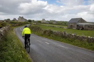 Cyclist in yellow reflective jacket riding along a damp country road in La Hague, bordered by green fields and stone walls, with cloudy sky and small hamlets in the background.
