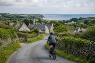 Cyclist on a quiet rural road in the Norman countryside, surrounded by stone walls and green fields with a distant view of the sea.