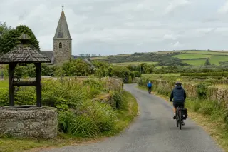 Cyclists riding on a winding rural road in La Hague, bordered by hedges, stone walls, and an old church, under a soft cloudy sky.