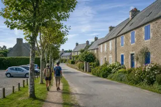 Deux personnes marchent sur un chemin à travers un hameau normand paisible, maisons en pierre et végétation fleurie sous un ciel clair.