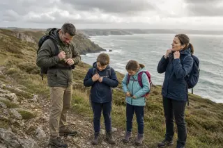 Famille en vêtements de randonnée sur un sentier côtier herbeux, prêts à marcher avec vue sur la mer et les falaises du Cotentin, dans une lumière naturelle avec ciel nuageux.