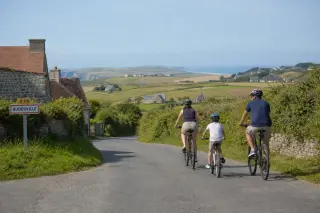 Family cycling on a small country road leaving a Norman village, passing green fields and hedgerows under a clear sky, with the open coast visible in the distance.