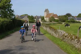 Family riding bicycles together on a quiet rural road surrounded by green fields and hedgerows near Branville-Hague, with a small church in the background on a clear day.