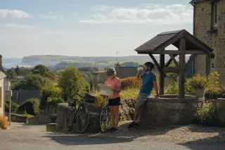 Two cyclists resting by a stone well in a Normandy village, with wildflowers, old buildings, and distant cliffs visible in soft morning light.