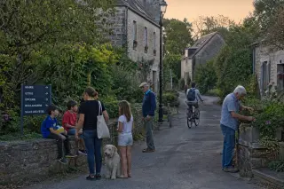 People of different ages interact naturally outside traditional stone houses in a Normandy hamlet at dusk, with subtle details showing local life and renewal, in soft, authentic light.