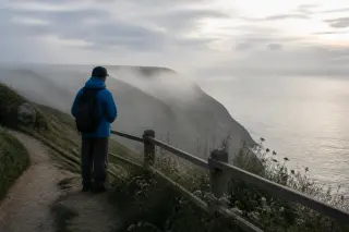 Solitary hiker facing away on a misty Cotentin cliff path at dawn, with diffused light and wild coastal scenery.
