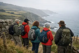 Group of hikers of mixed ages and backgrounds pausing on a coastal path in La Hague, looking out over cliffs and patchwork fields under a soft, overcast sky.