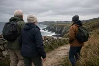 Three people of different ages walking along a rugged coastal path in the Hague, with cliffs, hedgerows and cloudy skies in a natural, candid atmosphere.