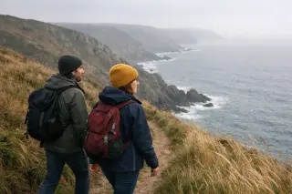 Two hikers walking naturally along the wind-swept cliffs of La Hague, with rugged coastline and misty sea in the background