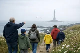 Family walking on a grassy coastal path near the misty Goury lighthouse, under a soft overcast sky, capturing an authentic and natural moment.