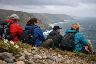 Several hikers rest by windswept cliffs near Jobourg, surrounded by wild coastal scenery under an overcast sky.