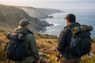 Two hikers overlooking coastal cliffs near Branville-Hague, pausing on a wild heath under soft morning light