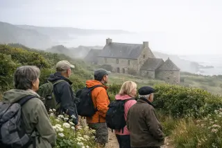 Group of hikers pausing near an old stone farmhouse on a coastal trail in La Hague, Normandy, surrounded by wild greenery and mist, with soft natural light.