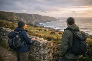 Two hikers in outdoor clothes pausing on a coastal path in La Hague, looking at cliffs and cows, with Norman stone walls and wild meadows at golden hour.