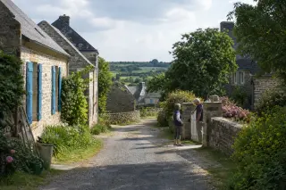 Hameau rural du Cotentin avec maisons en pierre, ruelle calme, végétation, deux habitants discutant près d’un portail, ambiance paisible et authentique.