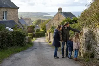 Normandy rural hamlet with stone houses, a family walking on a quiet lane, green hills and distant coastline under soft afternoon light.