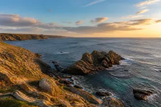 Open rocky coastline of Pointe de la Hague with dark rocks, blue-green sea, soft clouds, and golden sunlight highlighting the land at sunset