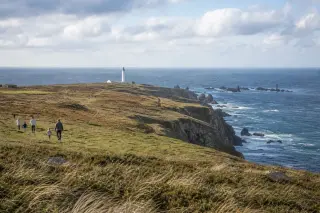 Panorama de la Pointe de la Hague avec des falaises surplombant la mer, herbes au premier plan, phare de Goury, promeneurs loin du bord et ciel lumineux