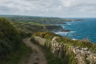 Panorama photoréaliste de la Hague avec falaises côtières, sentier bordé de haies et murets, mer agitée et petit village au loin sous ciel nuageux.