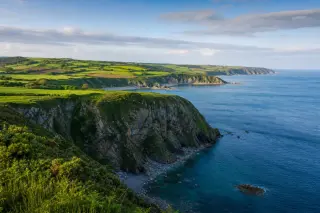 Panoramic coastal landscape of La Hague at sunrise, featuring cliffs, green fields, hedgerows, and a calm blue sea under a soft morning sky.