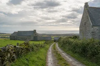 paysage rural de la Hague avec maison en pierre, haies, muret, chemins empierrés, champs, bâtiments agricoles, horizon côtier sous ciel nuageux