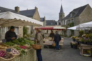 Petit marché de village dans le Cotentin avec stands de légumes, fromages et fleurs, quelques habitants discutant avec les producteurs, ambiance conviviale devant des maisons en pierre et un clocher.