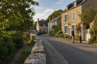 petite rue d’un hameau normand avec maisons de pierre rénovées, sentier entretenu, habitants en discussion et ambiance paisible de fin d’après-midi