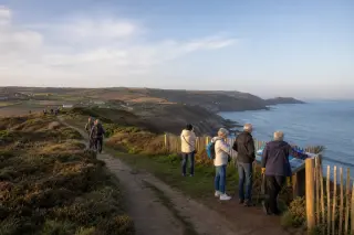 Promeneurs sur un sentier côtier balisé en Normandie, devant des landes, des falaises et un panneau d’information, avec arrière-plan rural et mer.