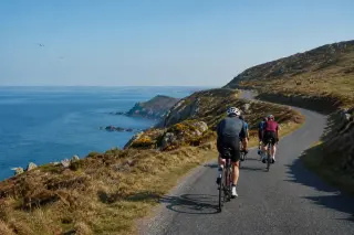 Route côtière sinueuse au bord de falaises, cyclistes avancent sous la lumière du matin avec la mer et des landes en arrière-plan, ambiance calme et naturelle.
