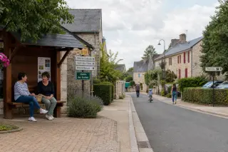 Rural hamlet with stone houses, safe sidewalks, subtle signs, greenery, and a few people of different ages using the shared public space.