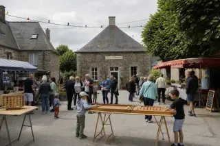 Scène vivante d’une fête de village normande sur une place entourée de maisons en pierre, stands associatifs, familles et enfants discutant et jouant sous un ciel nuageux.