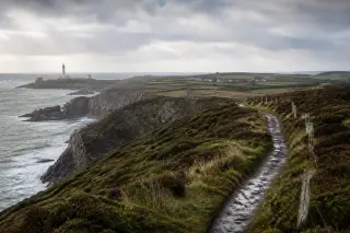 Sentier côtier étroit longeant une falaise normande à la Hague, vue sur la mer agitée, phare en arrière-plan, végétation basse, ciel nuageux, hameaux et prairies bocagères à distance.