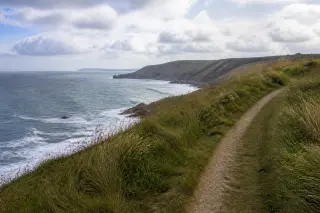 Sentier sur les falaises du Cotentin par temps lumineux et nuageux, herbe haute au vent, ciel changeant, vue calme sur la mer en contrebas.