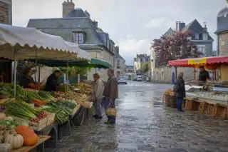 Small Normandy village market in the morning with a few stalls, people browsing for fresh produce, stone buildings in the background, and soft daylight.