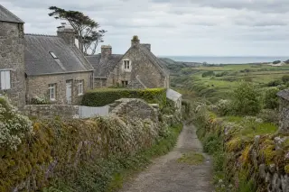 Un hameau rural en pierre dans la campagne du Cotentin, entouré de murets et de haies, avec vue sur des champs et la mer distante sous un ciel nuageux.