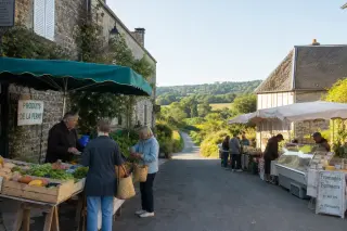 Village market in Normandy with people buying local produce at simple stalls along a quiet road bordered by stone houses and green hedgerows on a clear morning.